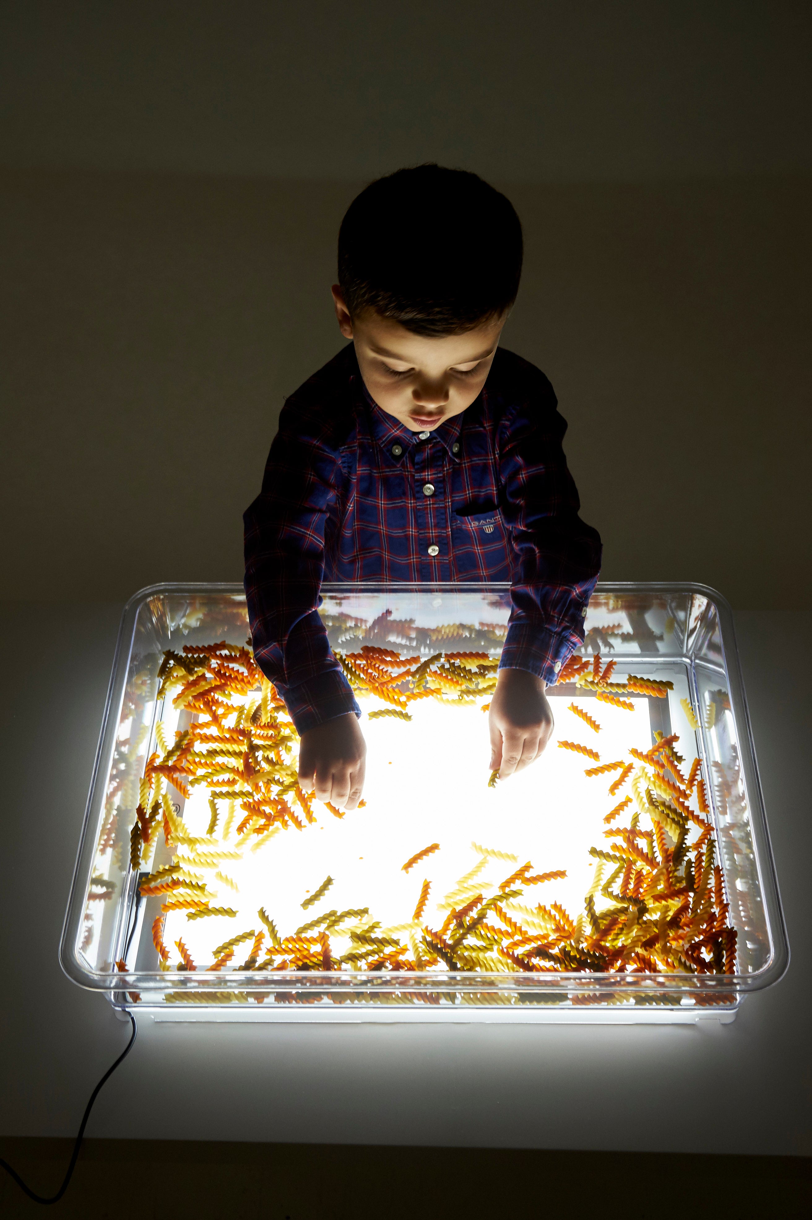 Rectangular clear exploration tray on a glowing light panel, child spreading golden glitter