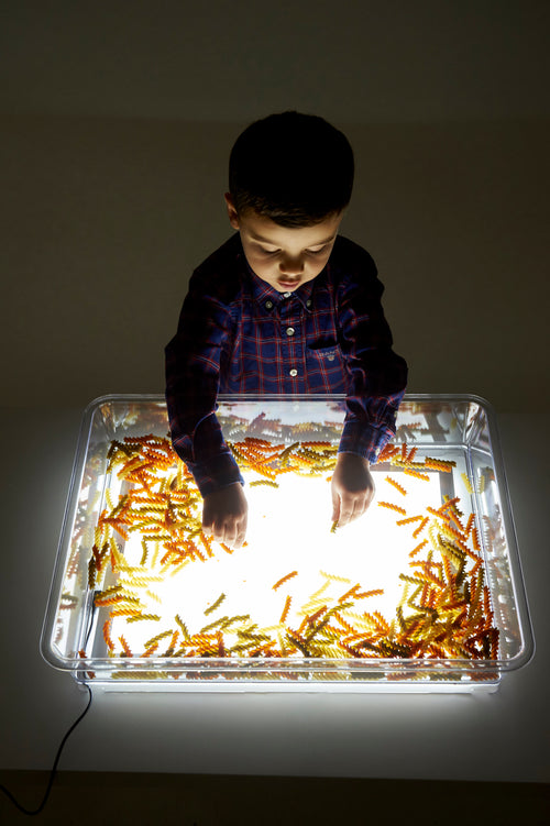 Rectangular clear exploration tray on a glowing light panel, child spreading golden glitter