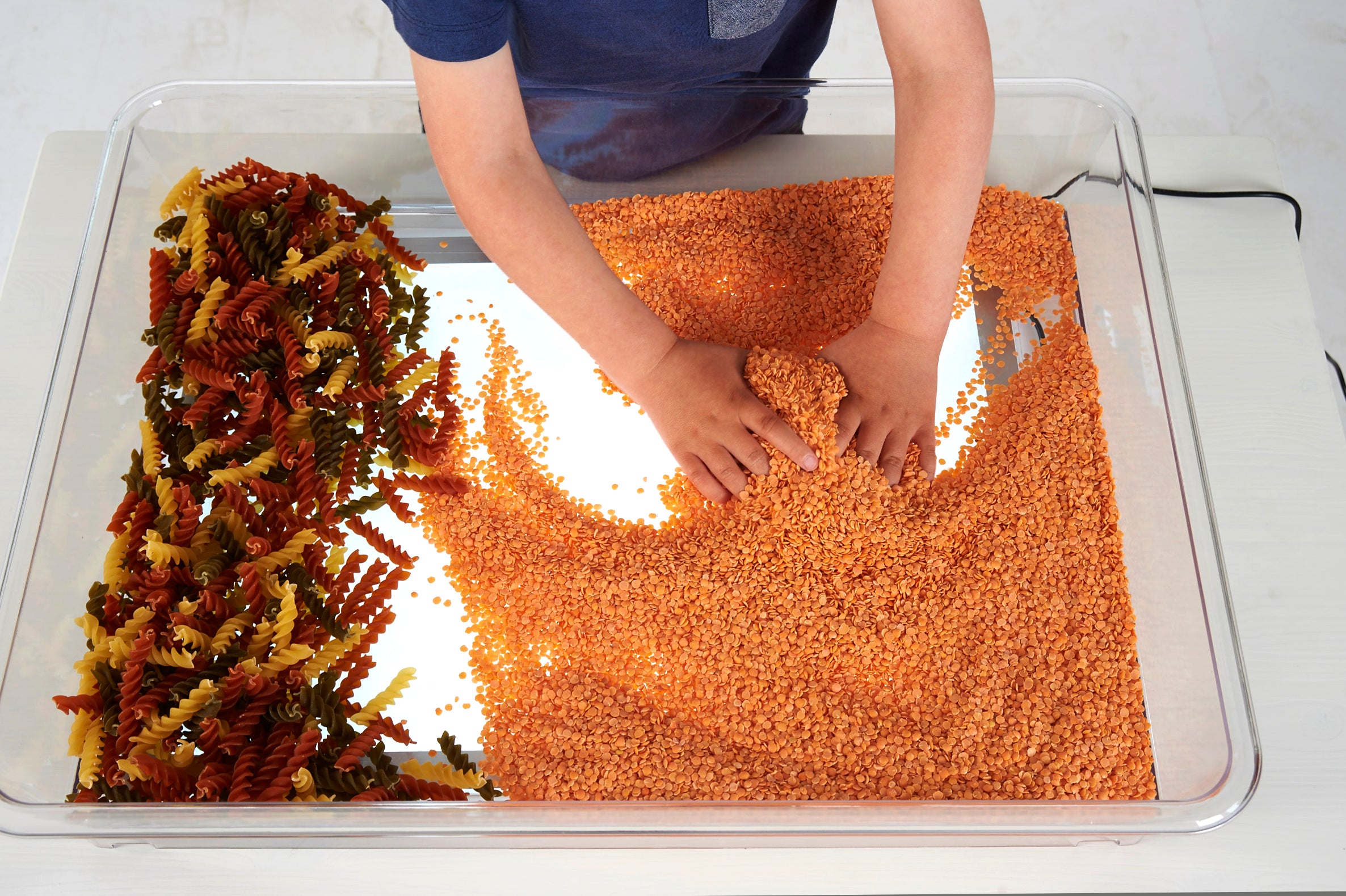 Clear plastic tray on a light panel, hands covering the surface with orange beads
