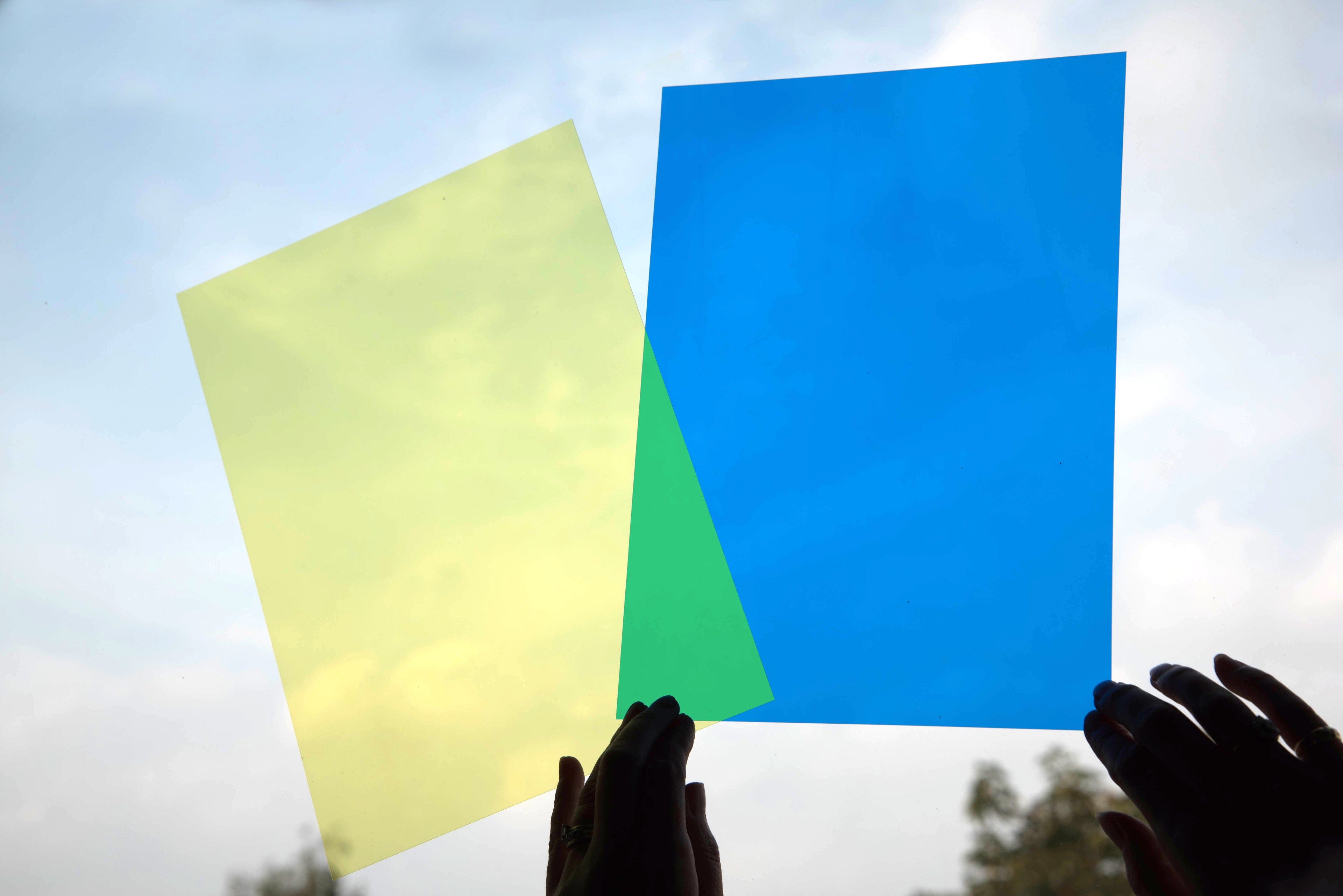 Colored acetate sheets outdoors held up by hands, with blue and light green panels
