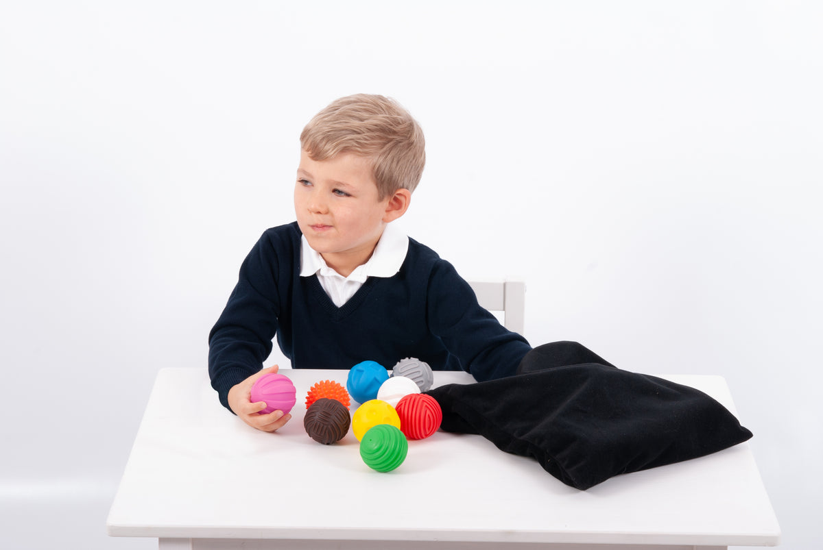 Child holding a pink textured ball with other colorful balls and black pouch nearby