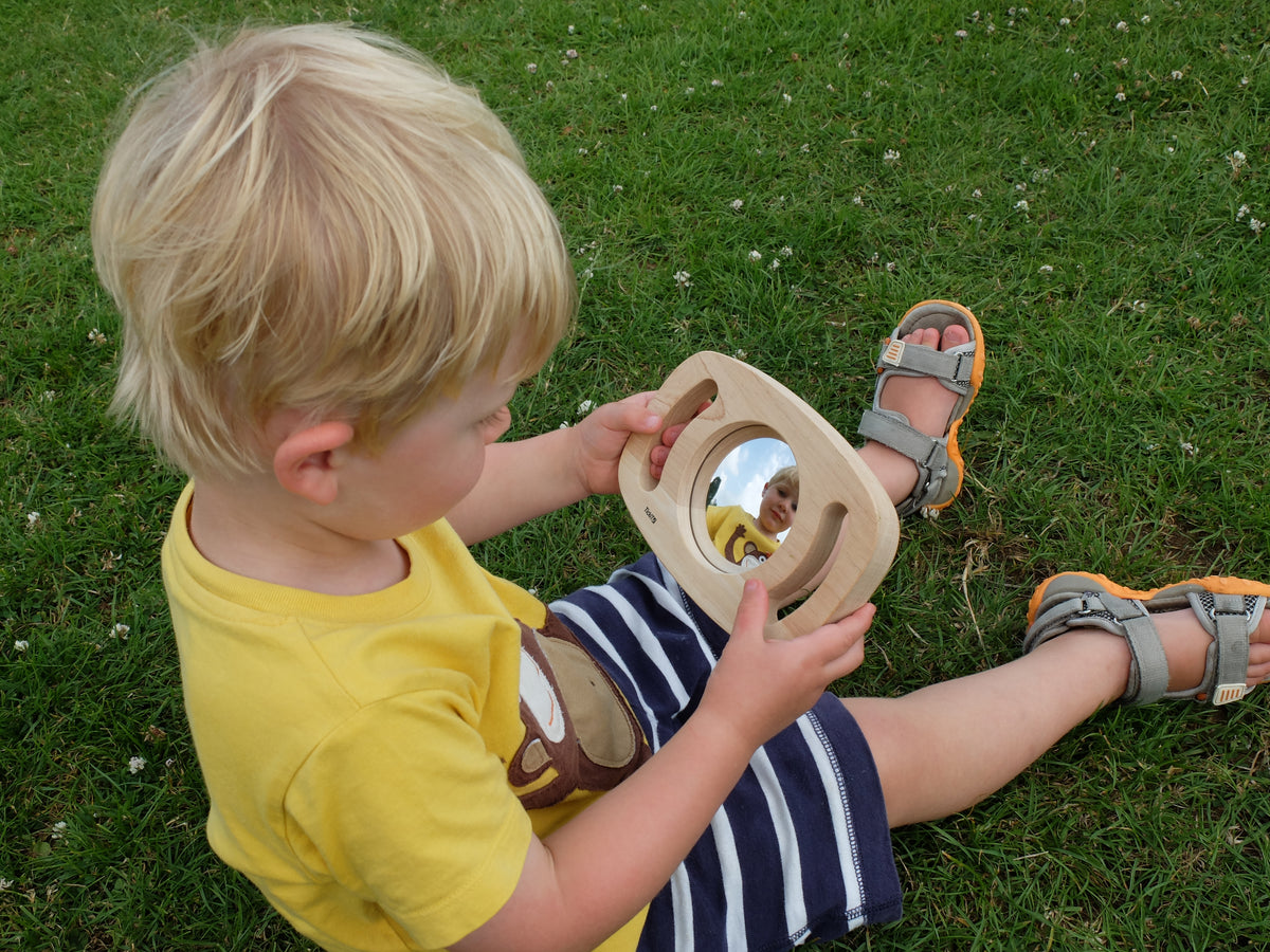Toddler holding an oval wooden frame mirror outdoors, mirror facing a child