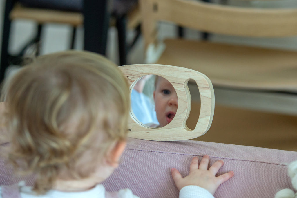 Oval wooden frame mirror with rounded grips on a surface, toddler view through