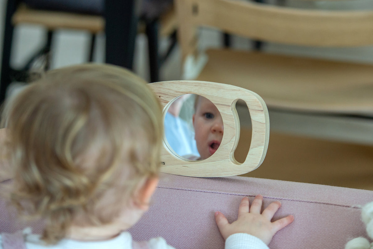Oval wooden frame mirror with rounded grips on a surface, toddler view through