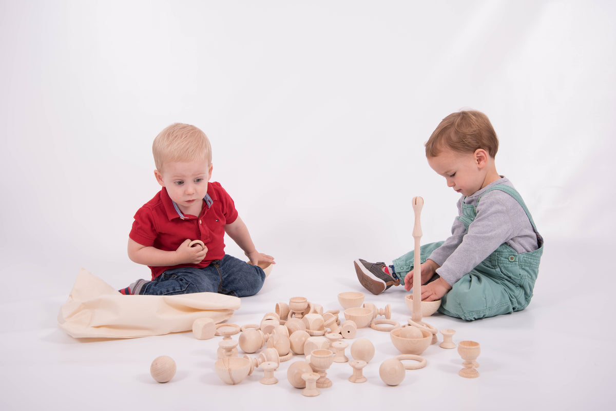 Wooden play starter set with many carved wooden pieces; two toddlers seated and building