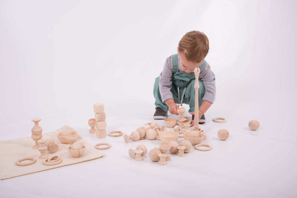 Wooden heuristic play starter set with towers, rings, and balls; toddler stands over pieces