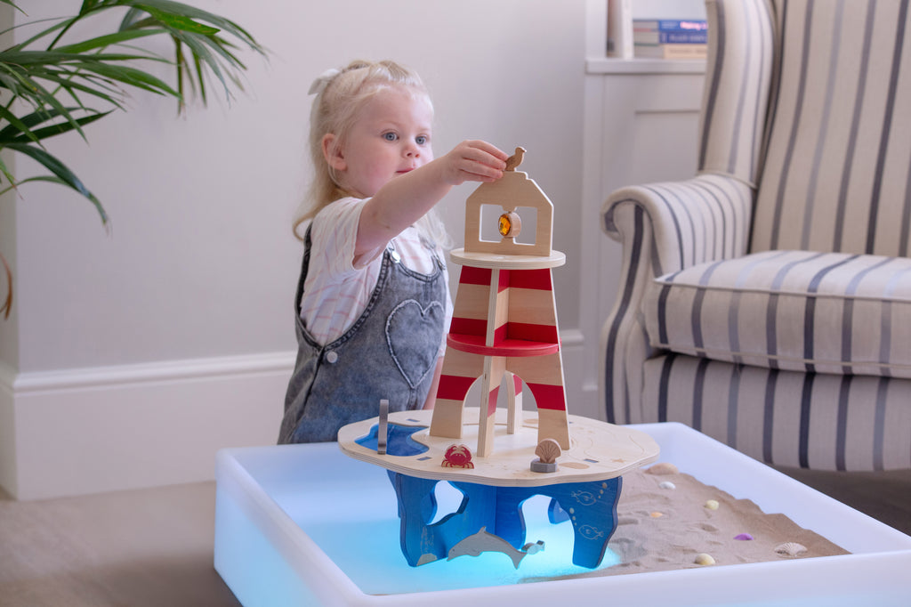 Wooden lighthouse playset on glowing water tray; child holds a small figure by the tower