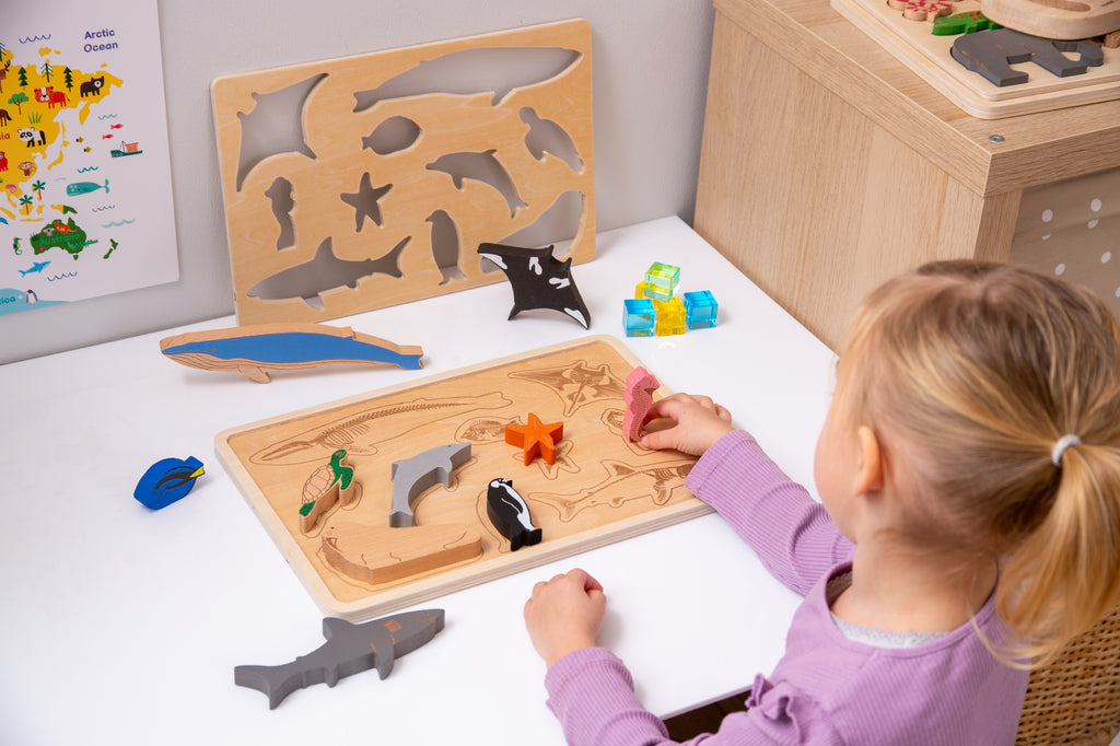 Child placing ocean animal puzzle pieces on two wooden boards; layered sharks, fish, and starfish visible.