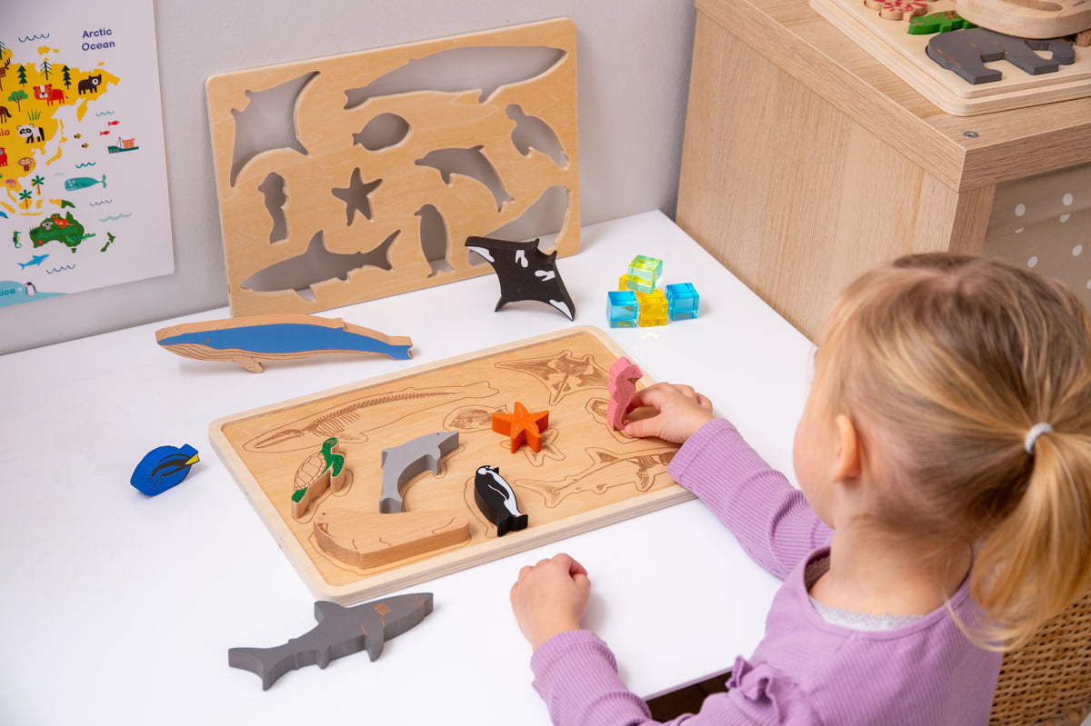 Child placing ocean animal puzzle pieces on two wooden boards; layered sharks, fish, and starfish visible.