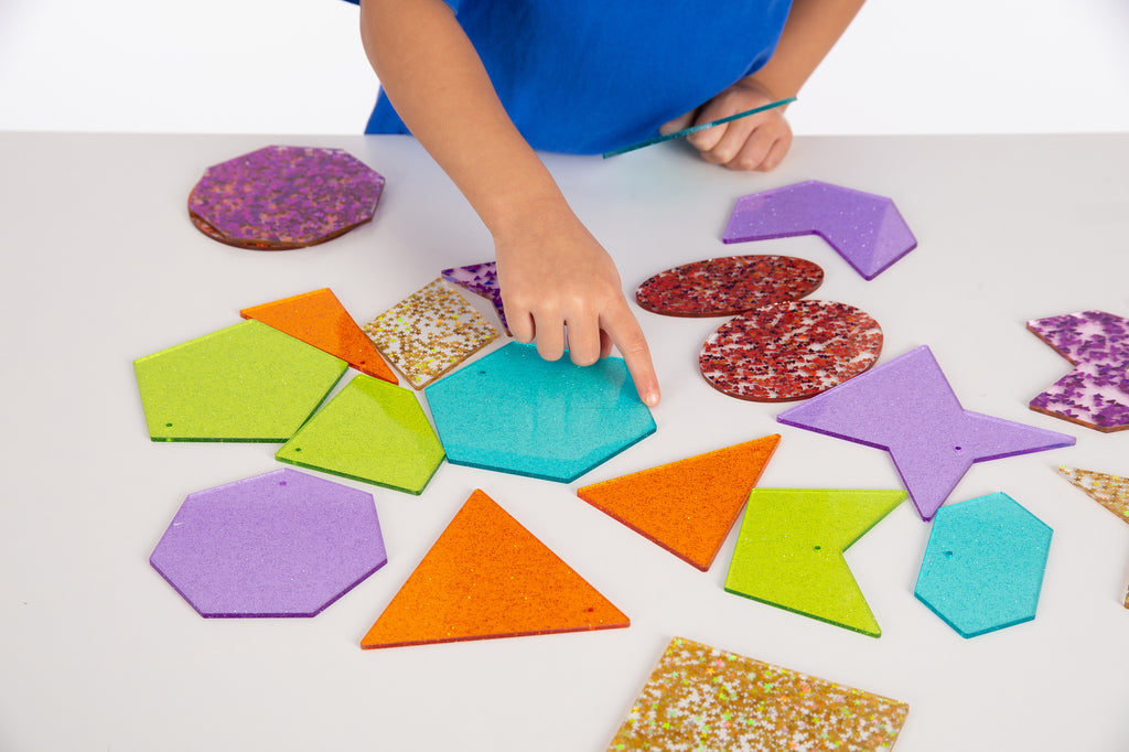 Hands arranging colorful glitter shapes, including stars, triangles, hexagons, and more