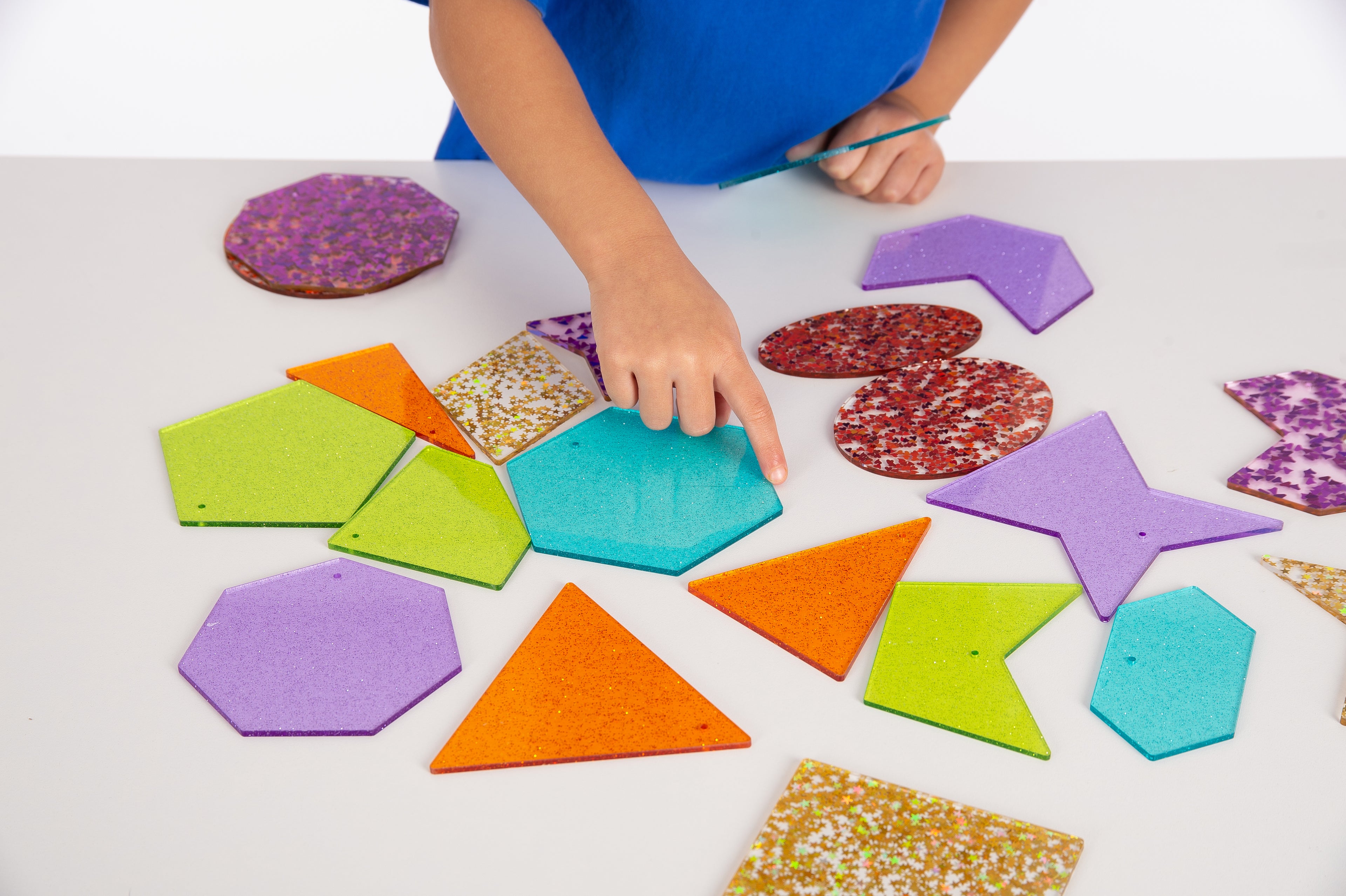 Hands arranging colorful glitter shapes, including stars, triangles, hexagons, and more