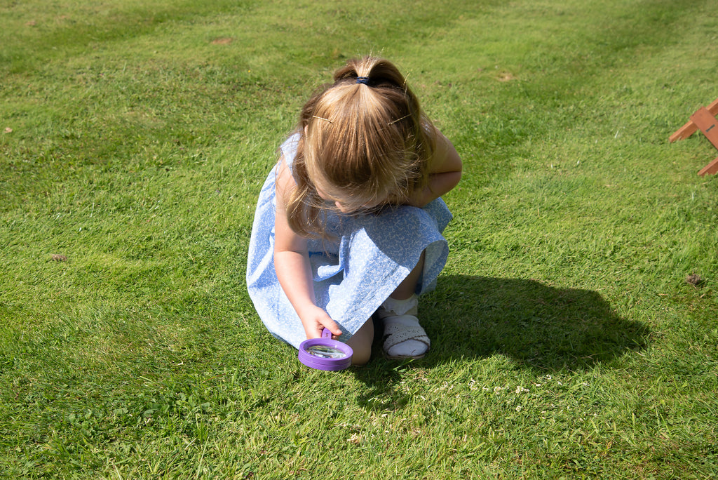 Girl crouching on grass holding a purple magnifying glass near the ground