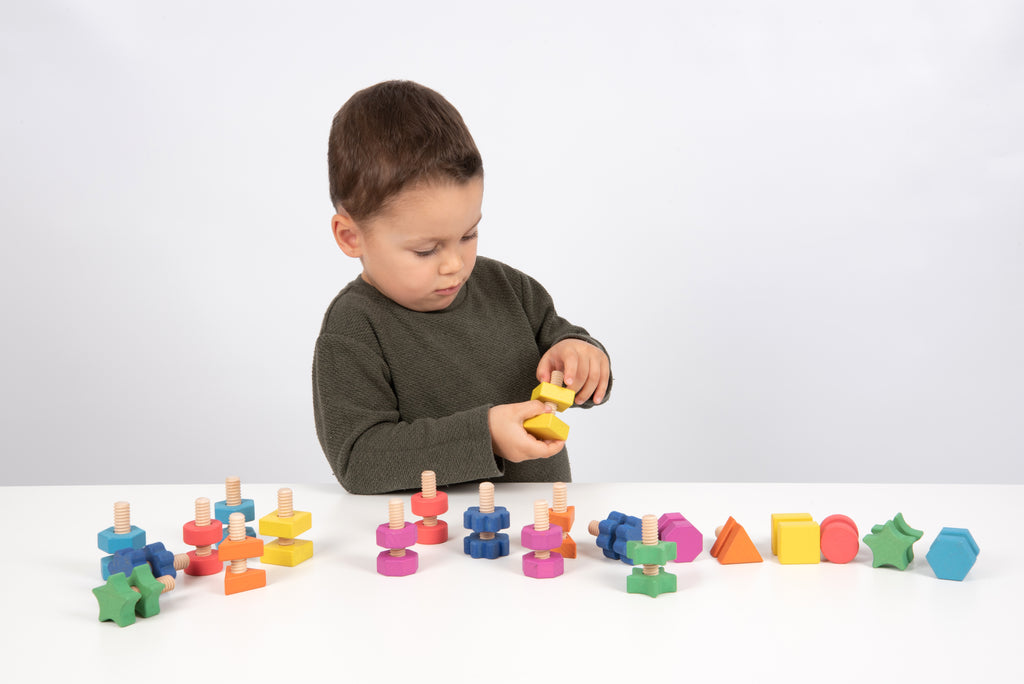 Child reaching for a yellow wooden nut-and-bolt piece among colorful pieces on table