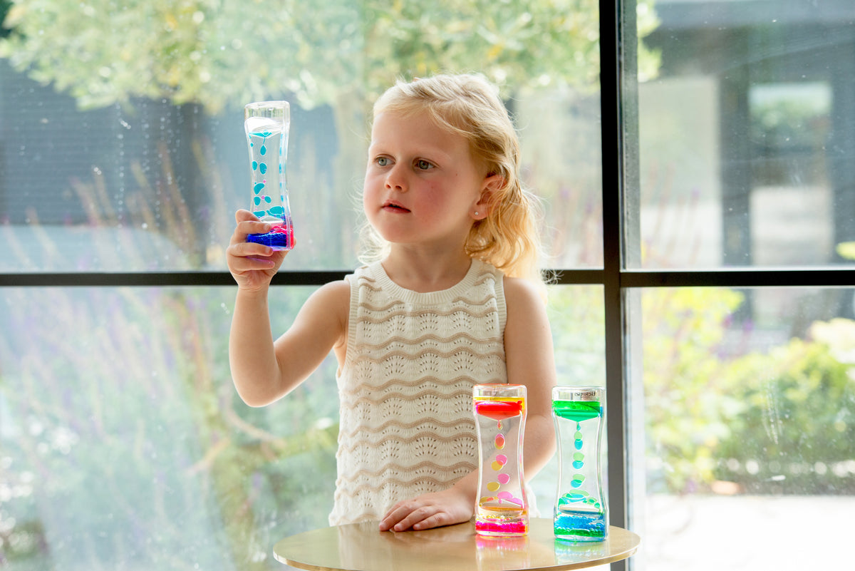 Child holding a blue rainbow sand timer; small colorful timers on clear tabletop