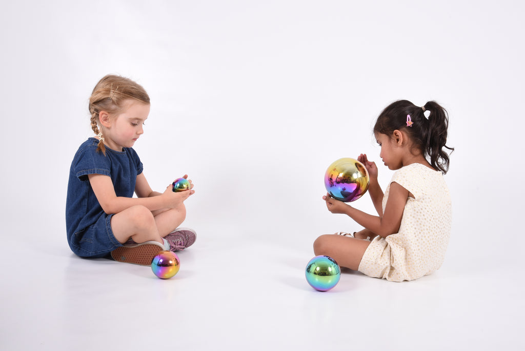 Set of four iridescent metallic sensory balls with rainbow sheen, two children sitting and playing