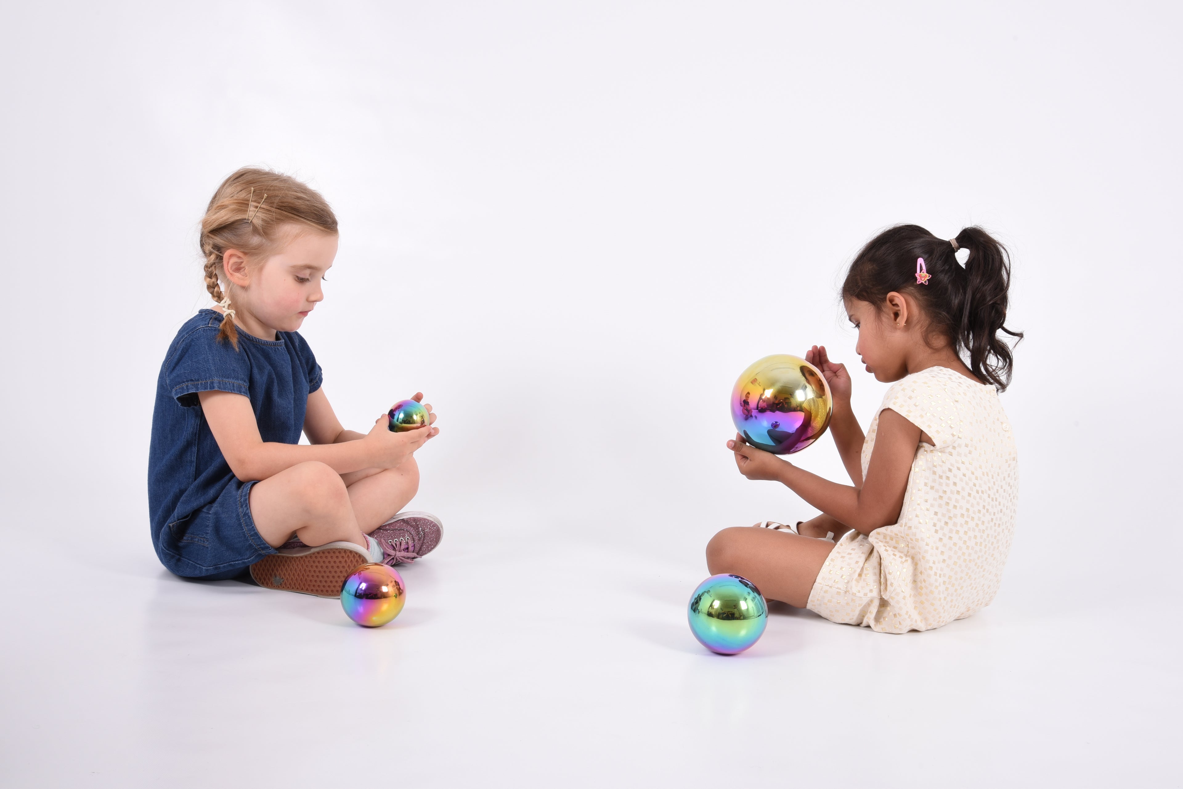 Set of four iridescent metallic sensory balls with rainbow sheen, two children sitting and playing