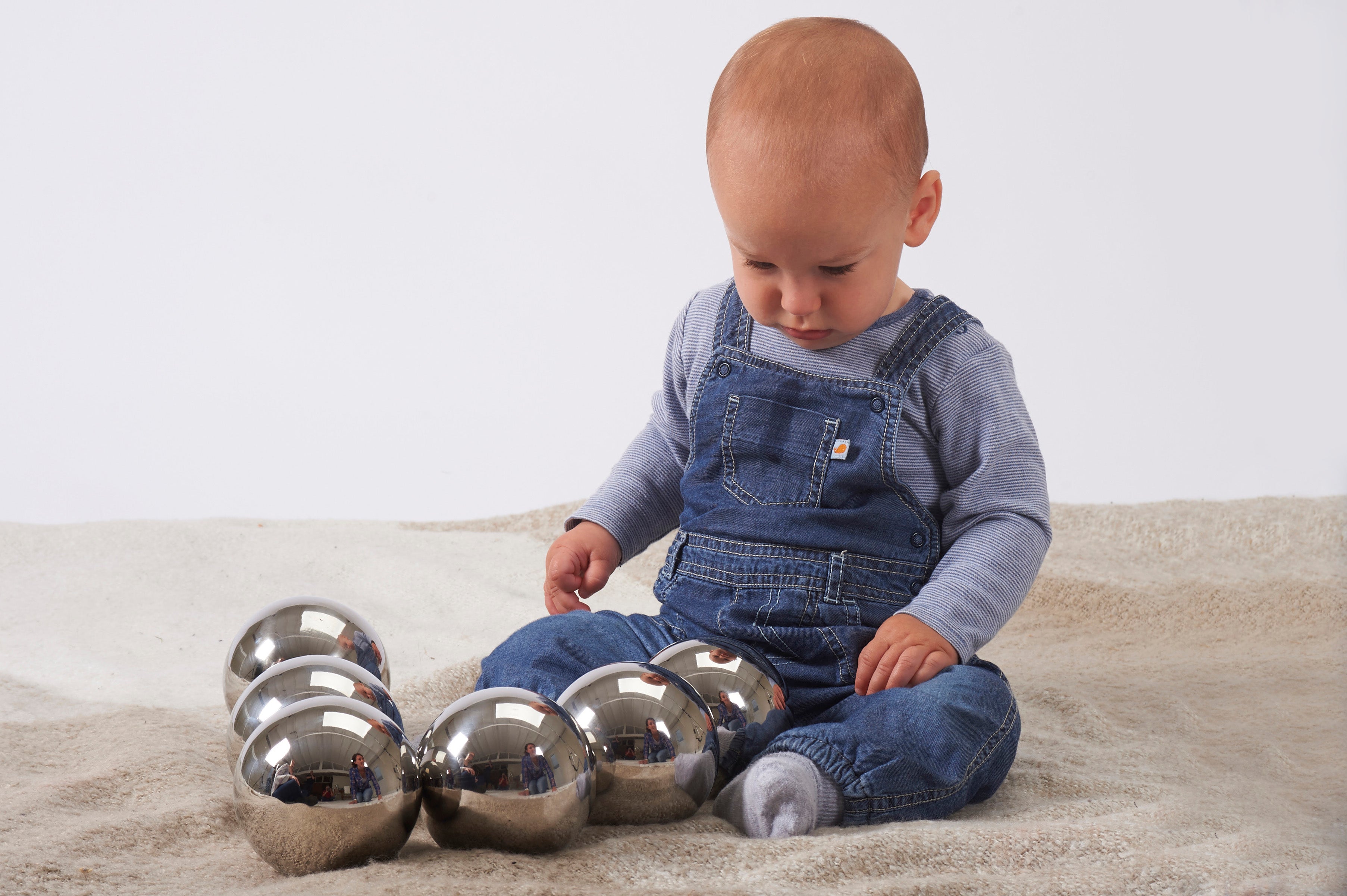 Set of six shiny steel sensory balls, child playing on beige fabric surface.