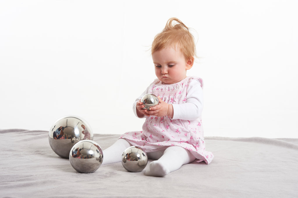 Set of shiny silver steel sensory balls, four sizes; baby holding one on a light blanket