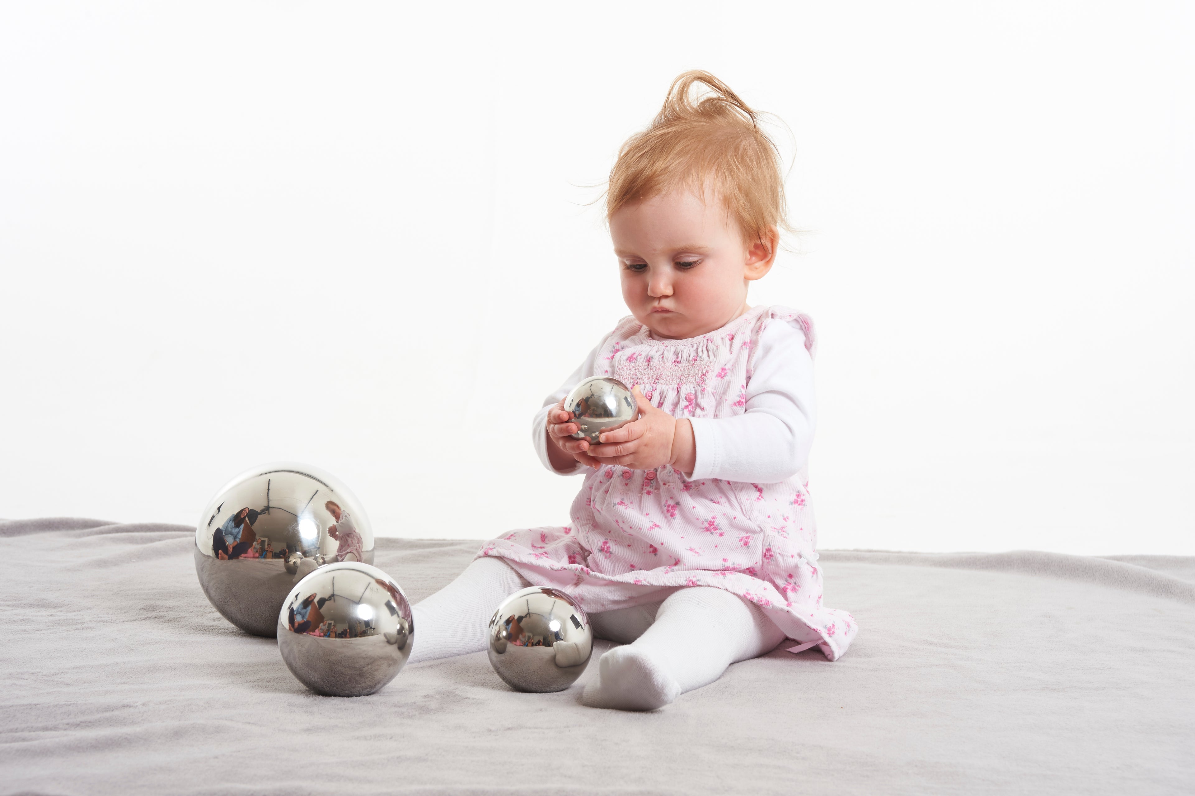 Set of shiny silver steel sensory balls, four sizes; baby holding one on a light blanket