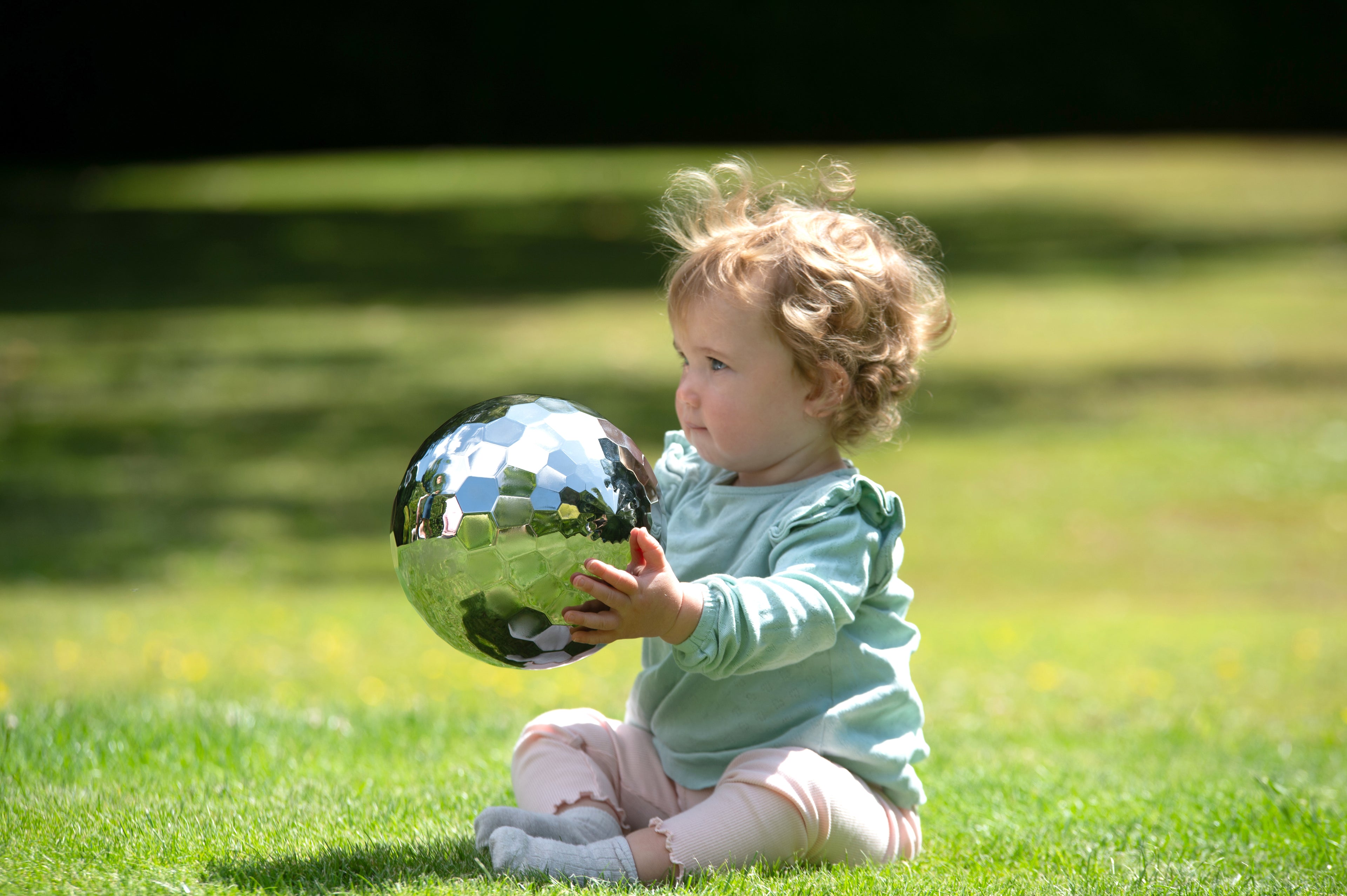 Steel tumble ball with mirrored facets held by a toddler in a grassy yard
