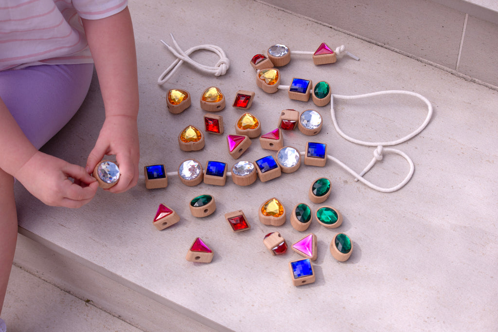 Necklace-in-progress of lacing jewels on cords: blue squares, pink triangles, green ovals, gold hearts