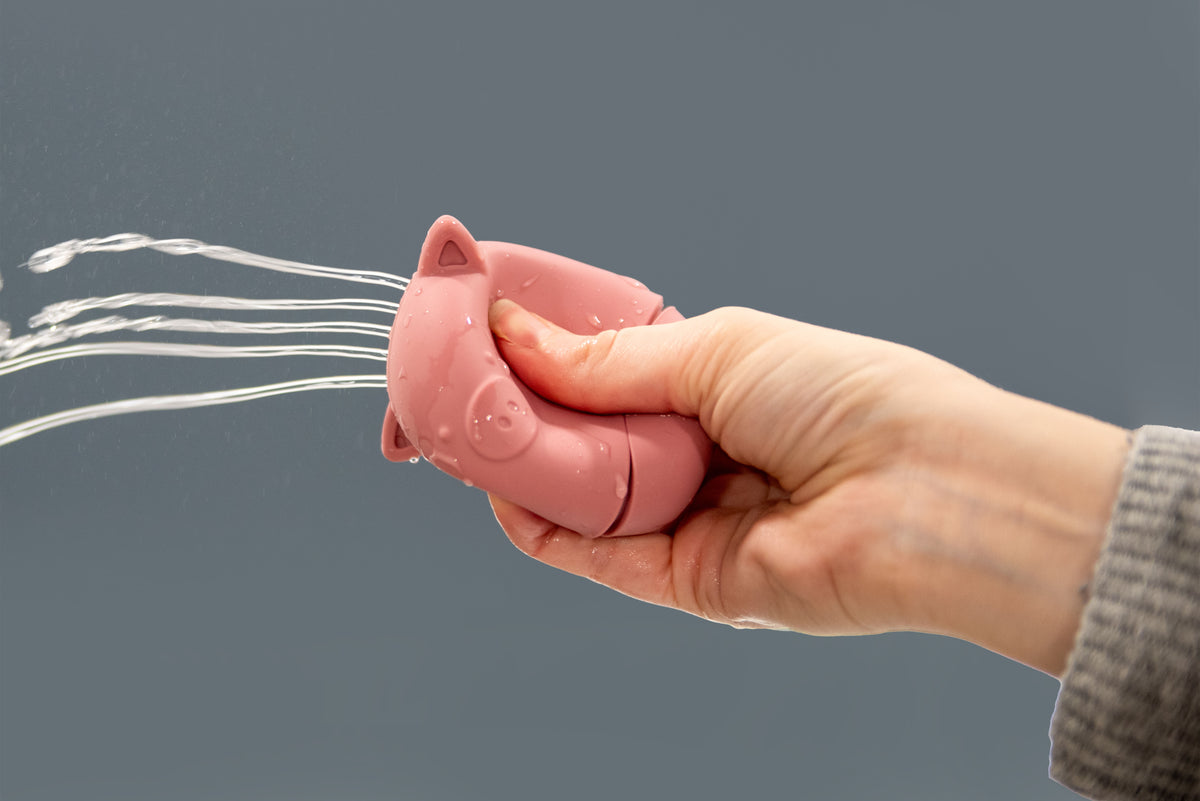 Pink bear-shaped silicone squirter toy held in hand with water stream.