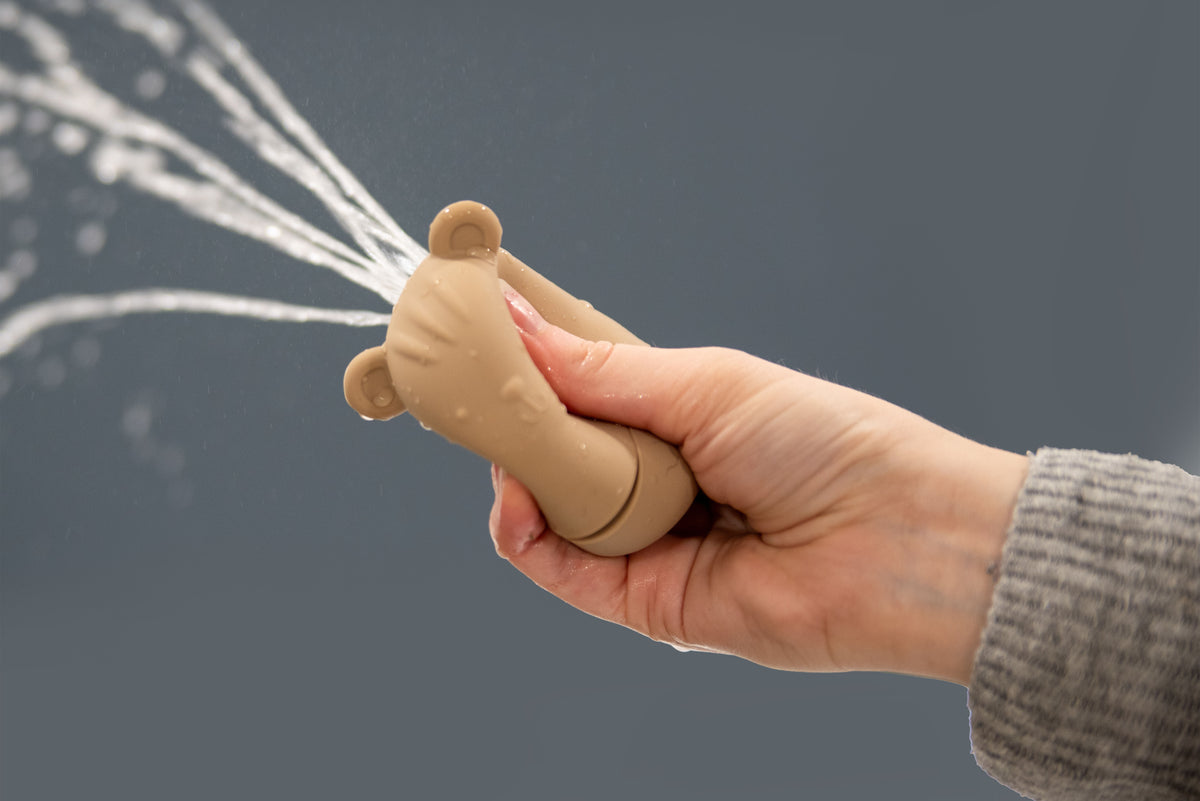 Beige bear-shaped silicone squirter toy held in hand with water stream.