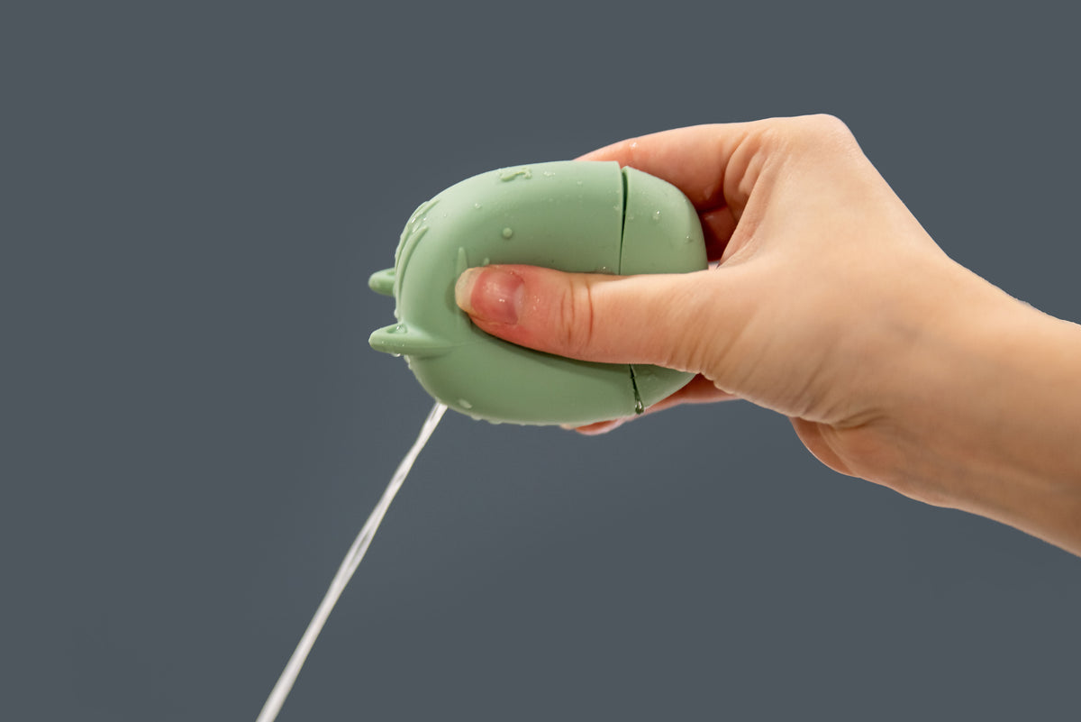 Green animal squirter toy with squeeze handle and spout, held in a hand