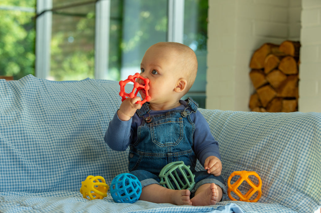 Rainbow flexi ball toy set; baby holds a red textured ball with other colors nearby