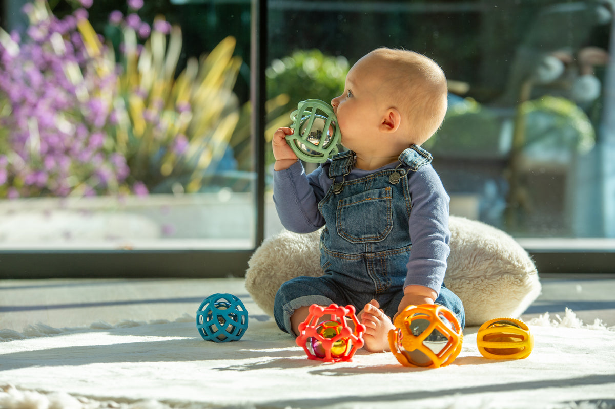 Rainbow flexi ball toy set; baby holds a green textured ball with other balls on floor