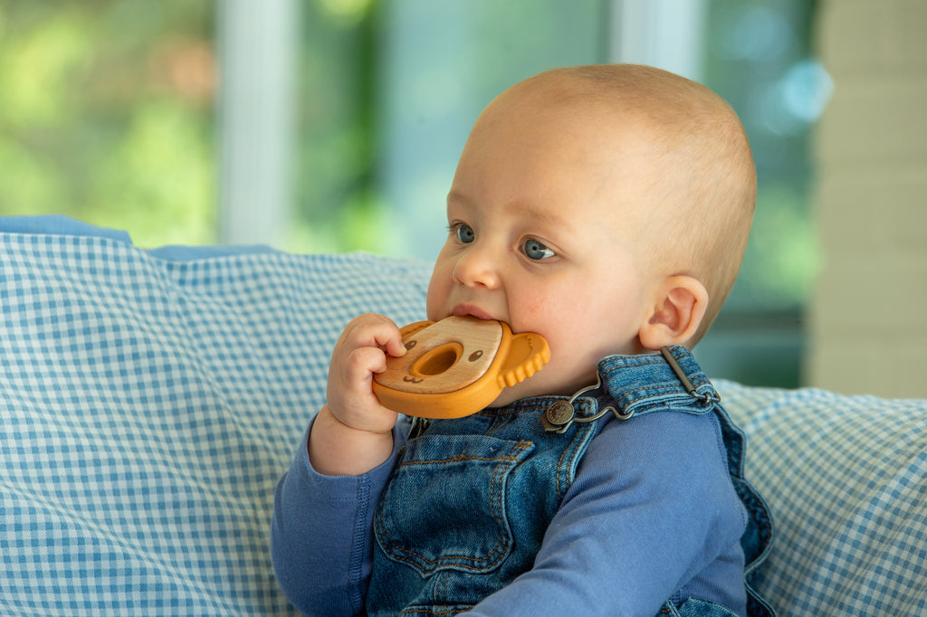 Baby chewing orange koala teether with round center ring while wearing blue denim