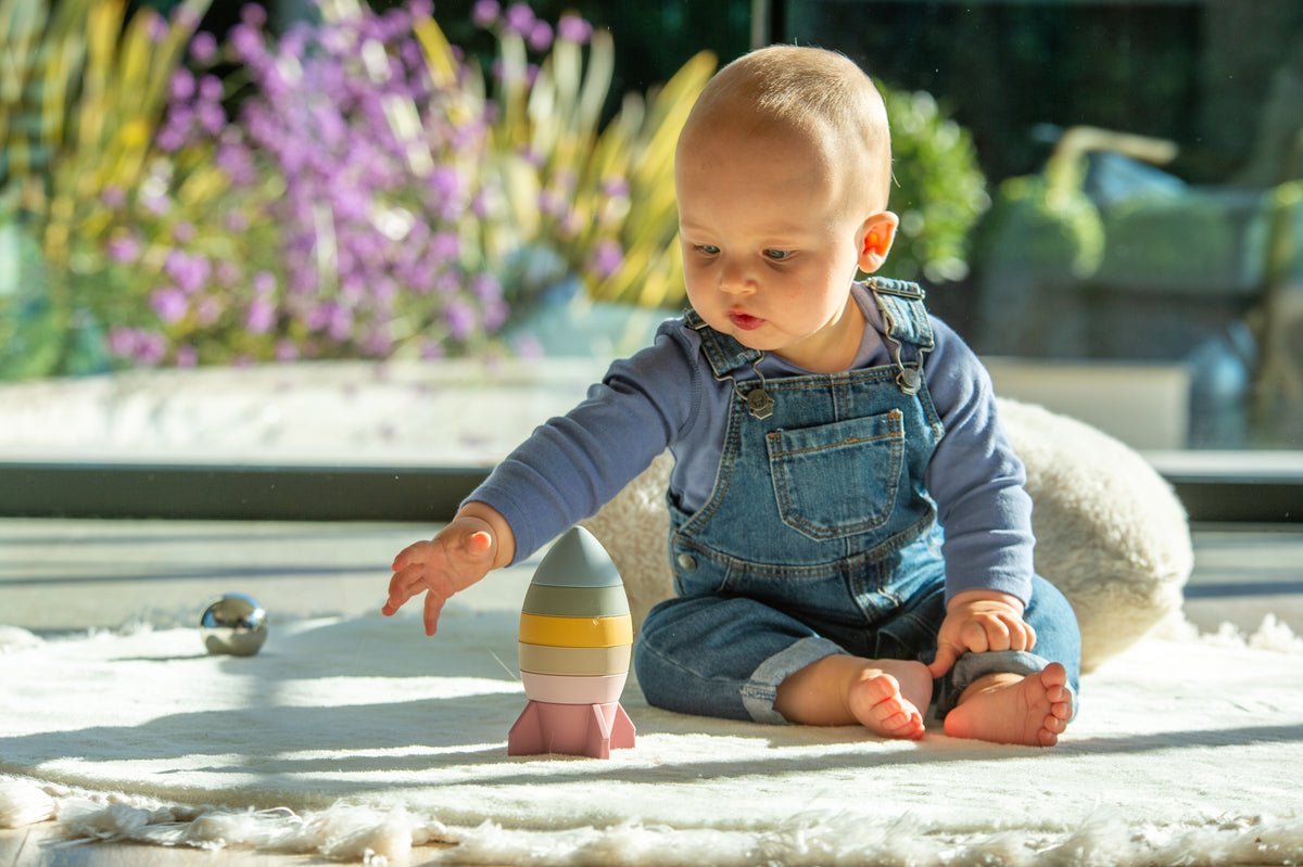 Baby reaching toward a rocket stacker toy on a plush outdoor mat
