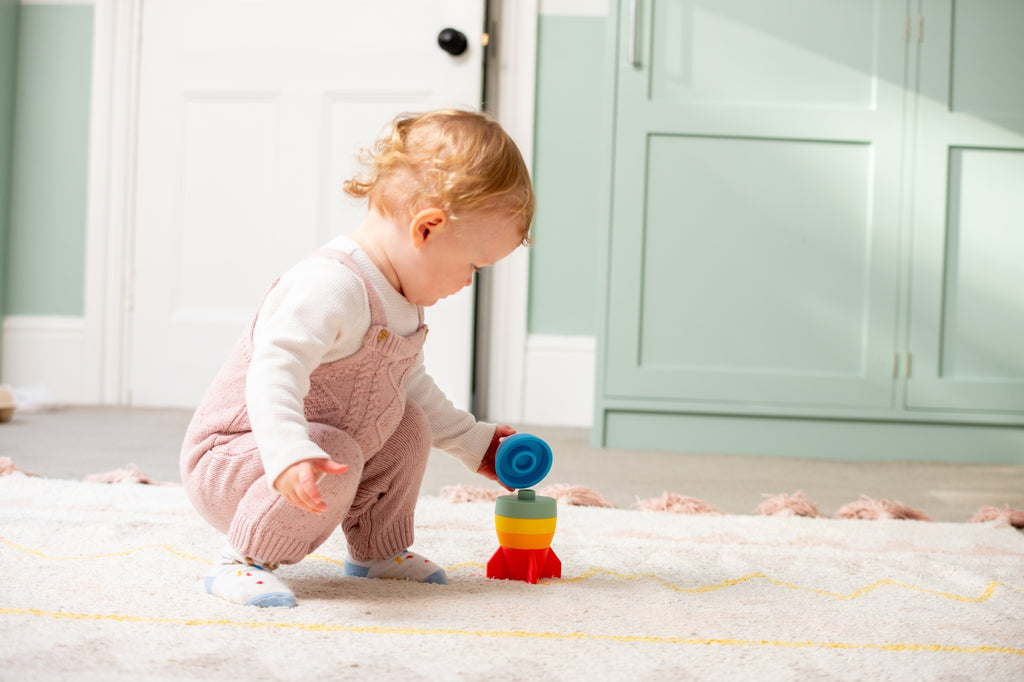 Child playing with rainbow rocket stacker toy on a soft play rug.