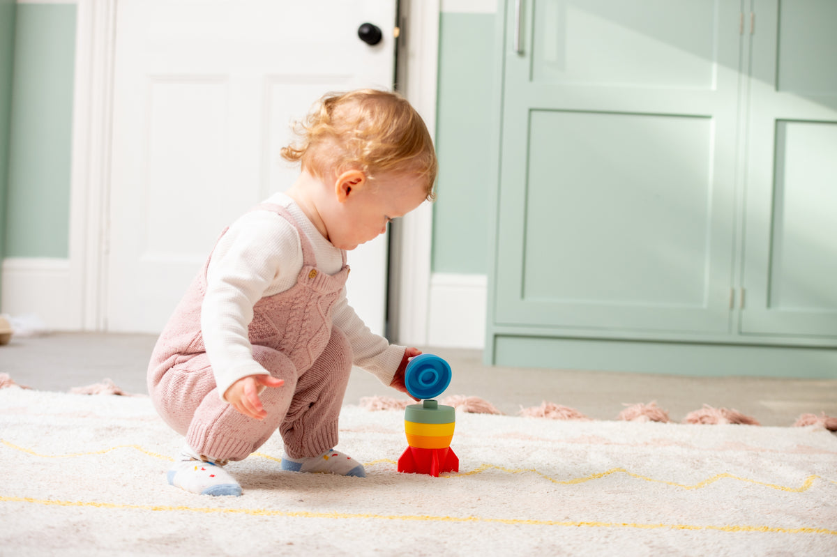 Child playing with rainbow rocket stacker toy on a soft play rug.