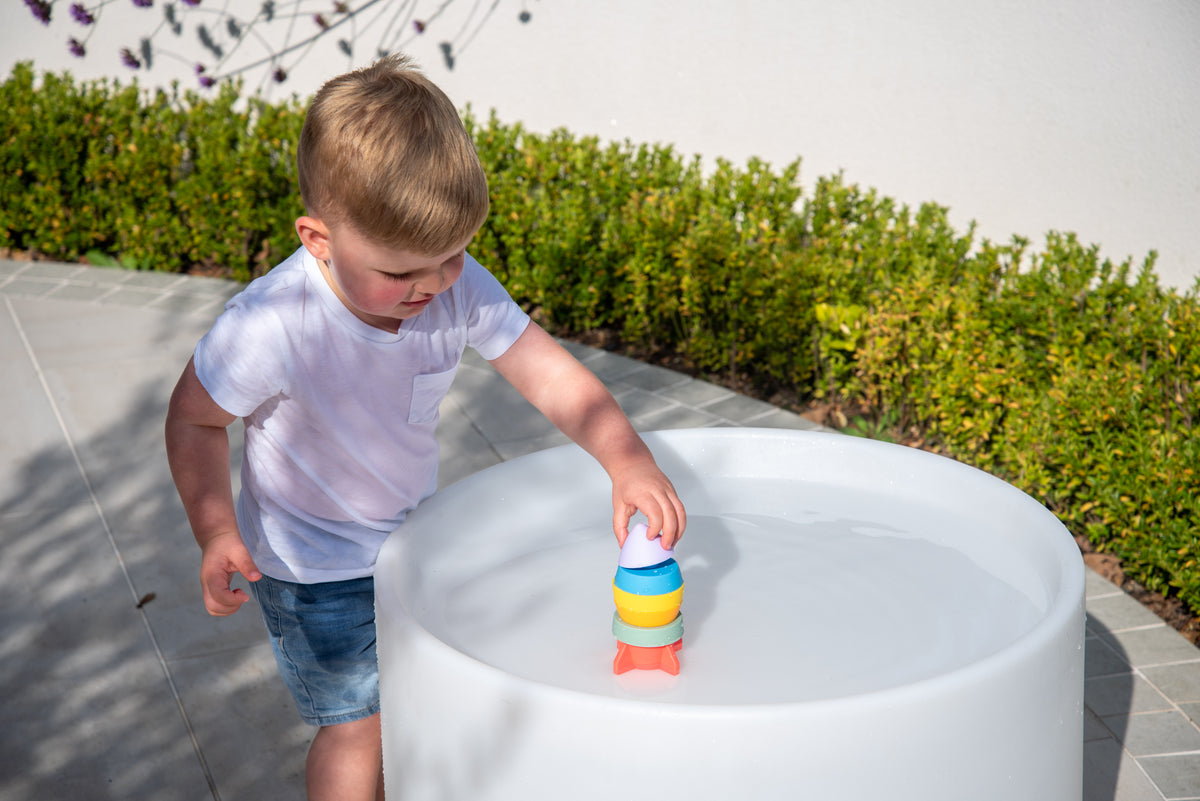 Rainbow rocket stacker toy in a child's hands beside a large white play tub.