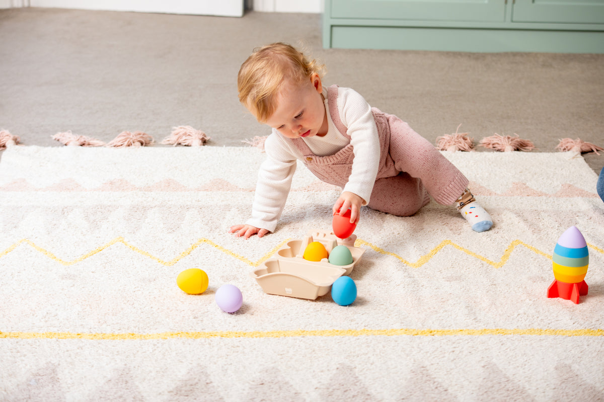 Toddler playing with colorful sensory eggs scattered on a rug and beige tray