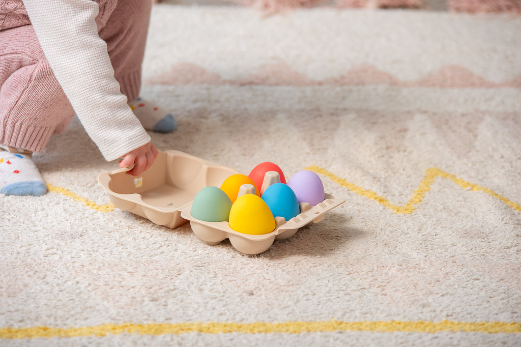 Hand placing rainbow sensory eggs into beige molded tray on a soft mat