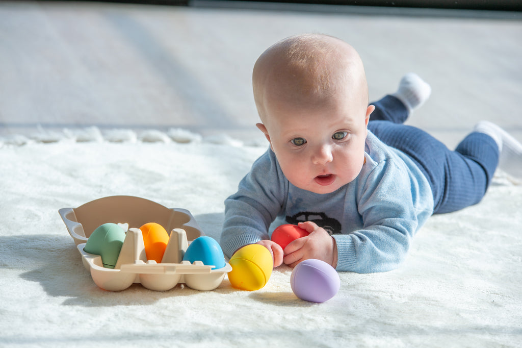 Baby on floor holding a purple sensory egg, with rainbow eggs in beige tray