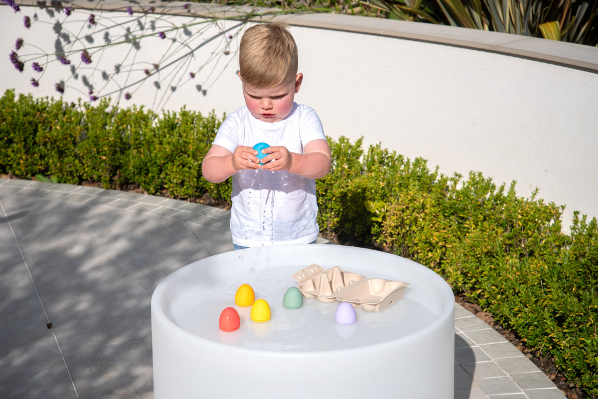 Child standing by round white sensory table with a few rainbow eggs