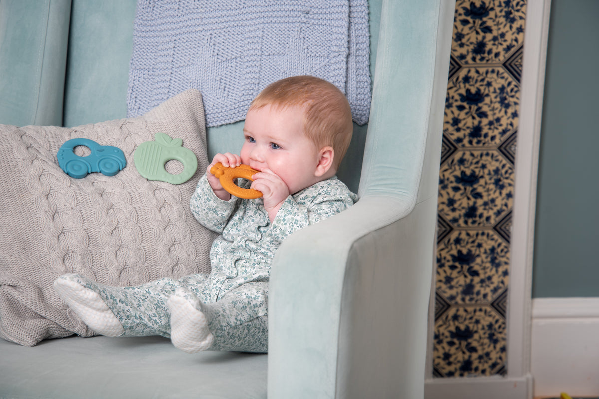 Baby holding orange silicone ring teether with green and blue toys on pillow
