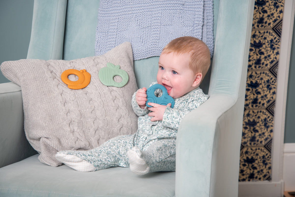 Baby holding blue silicone car-shaped teether in hands while lounging on couch
