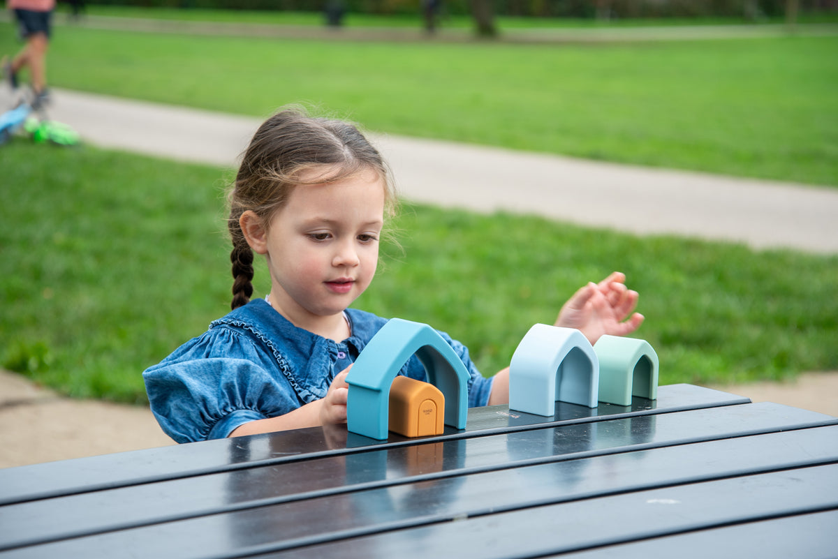 Child holding teal stacking house toy on outdoor bench, orange door visible