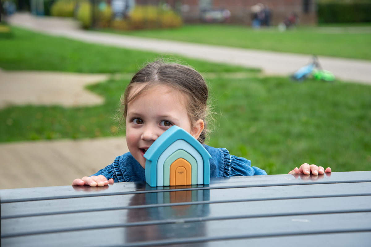 Small child behind bench holding teal stacking house toy with orange door