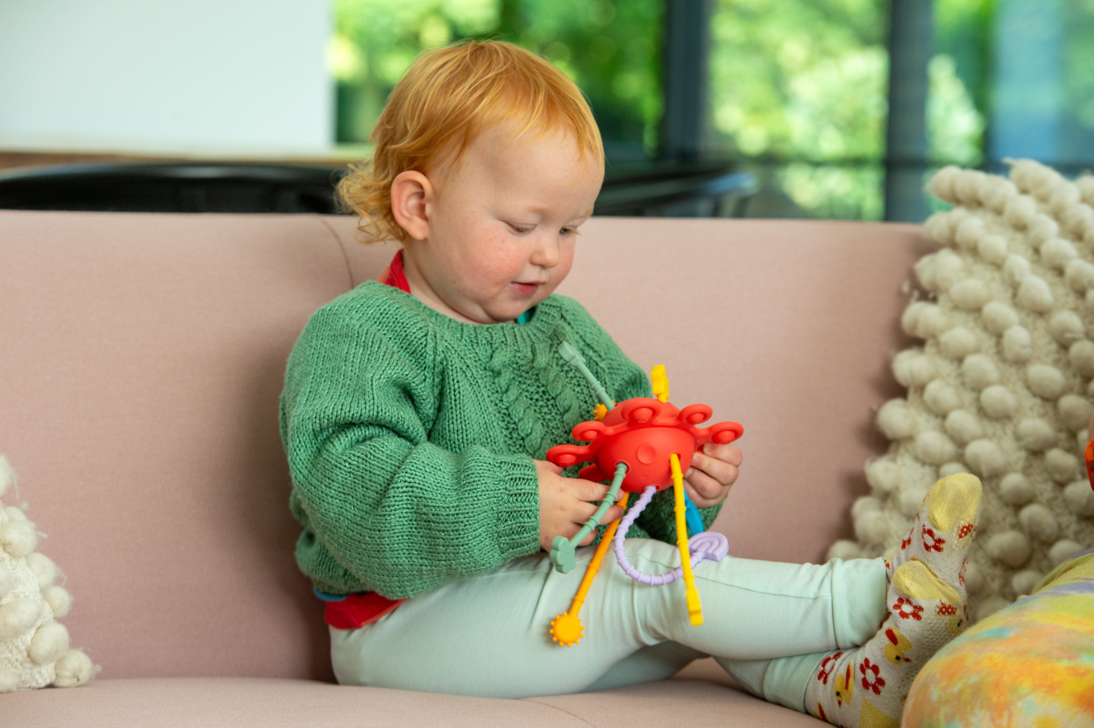 Toddler holding Silisoft rainbow zip pull toy with red center and dangling straps