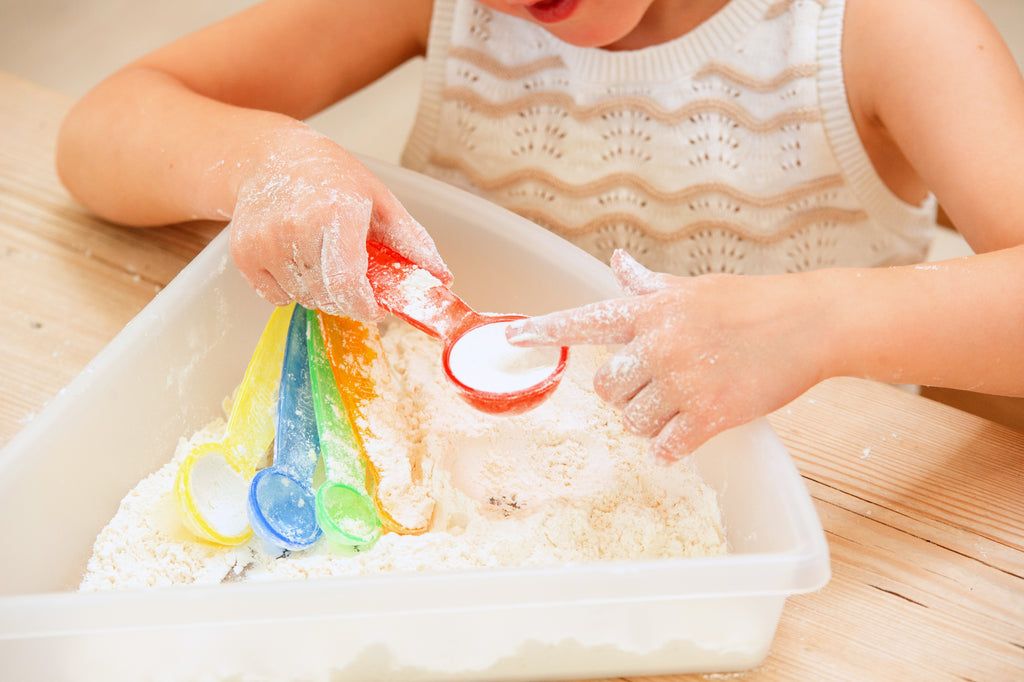 Translucent measuring spoons pouring colored powder into a plastic bin.