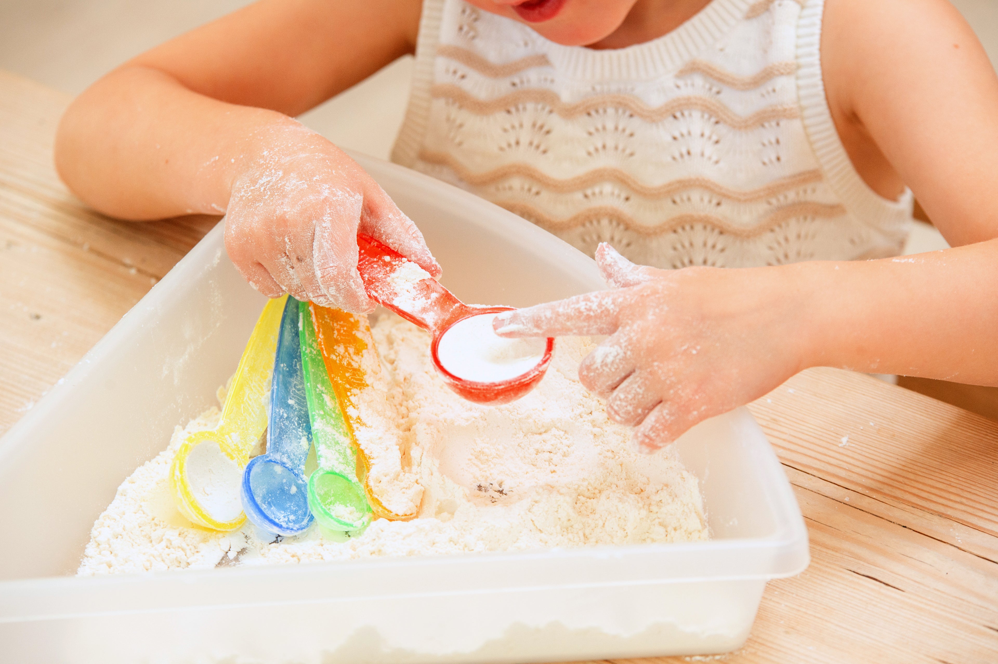Translucent measuring spoons pouring colored powder into a plastic bin.